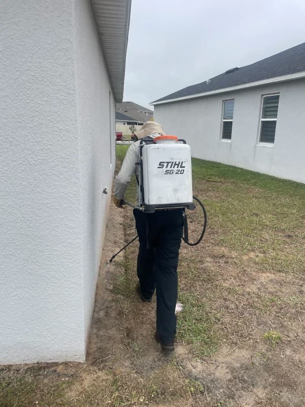 Crew member spraying weeds with a backpack sprayer along the side of a house